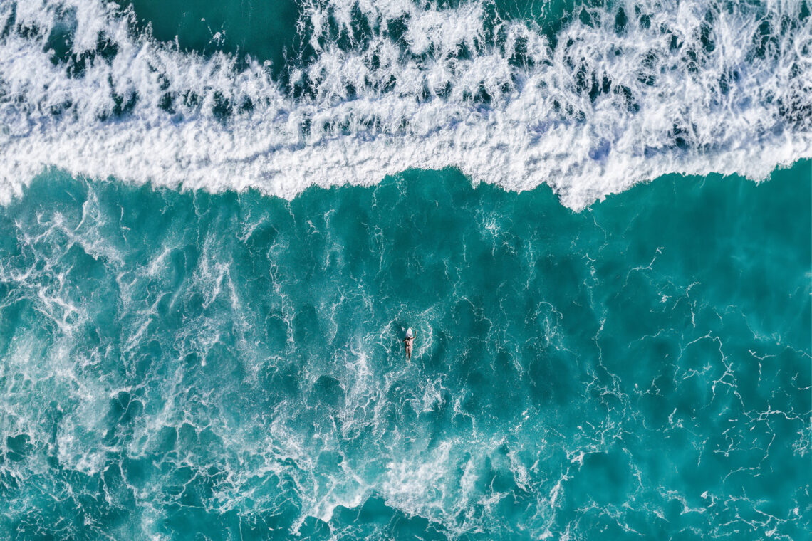 An arial view of a persons paddling into the vastness of the ocean on a surfboard.