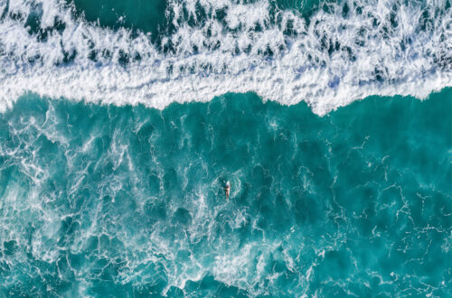 An arial view of a persons paddling into the vastness of the ocean on a surfboard.